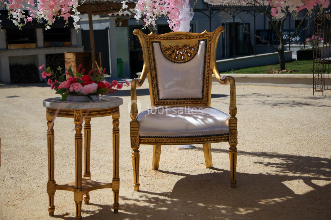 Fauteuil doré et table d'appoint ornée de fleurs roses, disposés en extérieur sous des décorations florales.