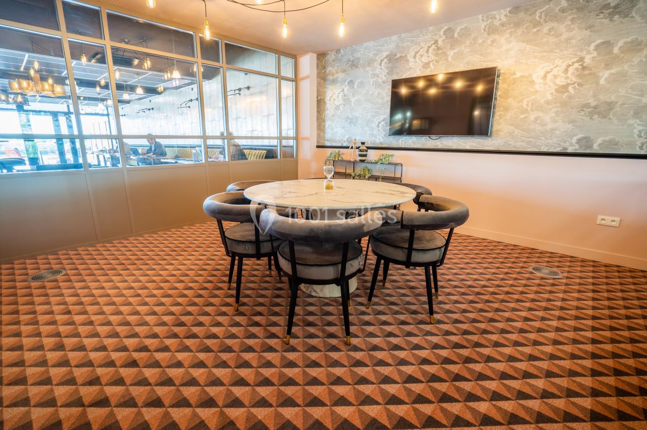 Salle de réunion avec table ronde, chaises rembourrées, écran mural et sol à motifs géométriques.