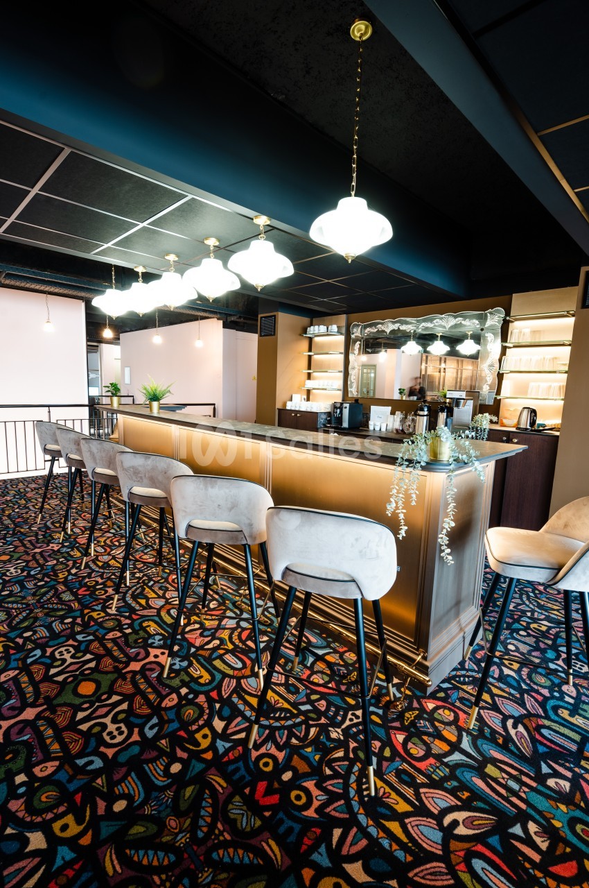 Comptoir de bar en bois clair avec tabourets, éclairé par des suspensions, sur un tapis coloré à motifs géométriques.