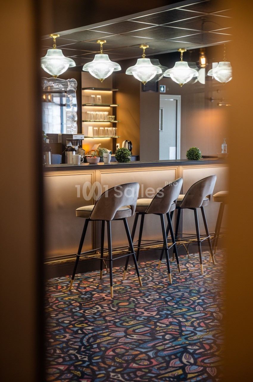 Bar élégant avec comptoir beige, tabourets rembourrés, suspensions lumineuses et sol à motifs colorés.