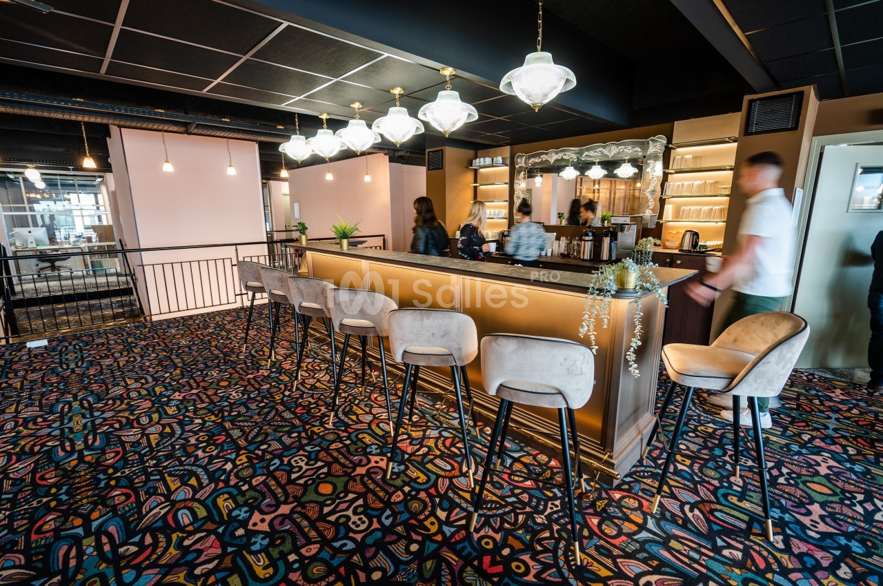 Bar moderne avec comptoir éclairé, tabourets rembourrés et tapis coloré, quelques personnes en arrière-plan.