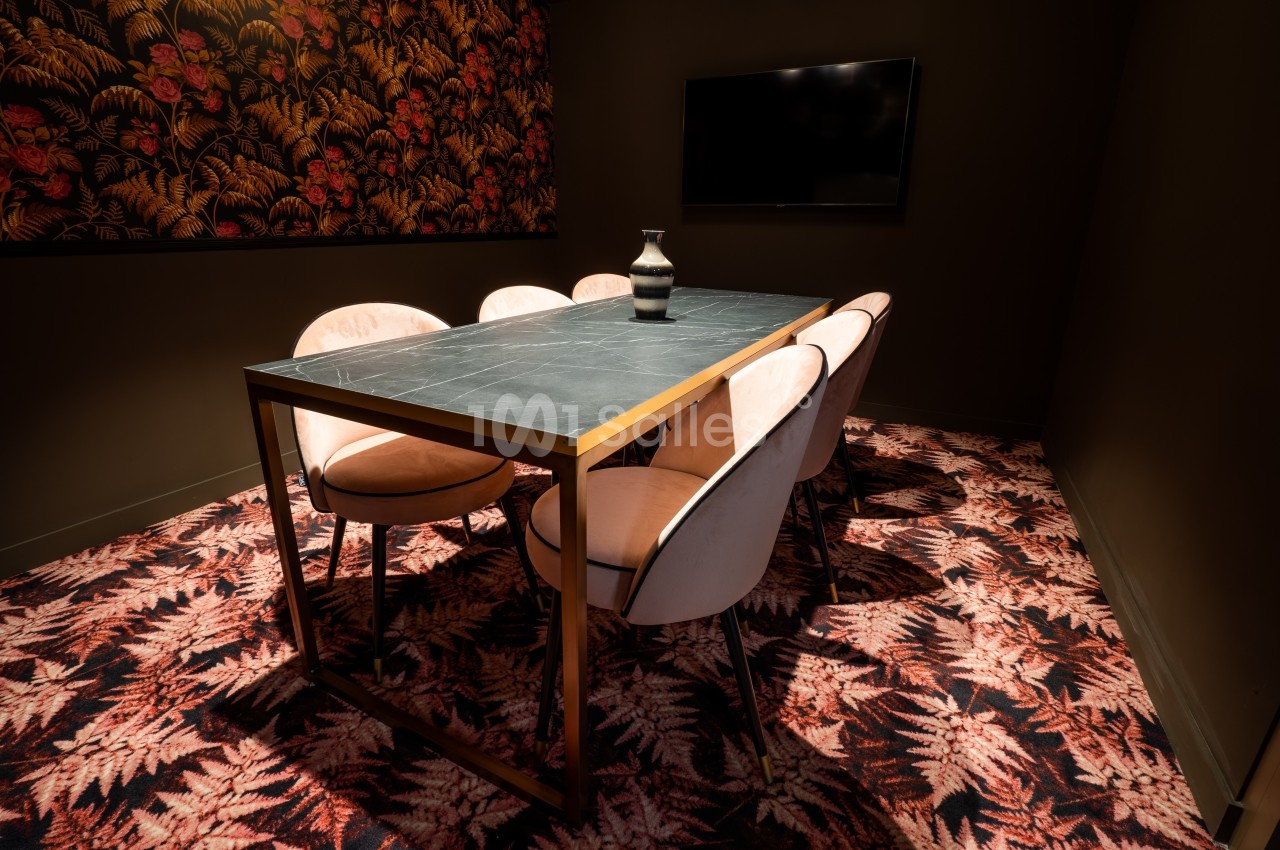 Table rectangulaire noire entourée de chaises roses dans une pièce sombre avec tapis à motifs végétaux et écran mural.