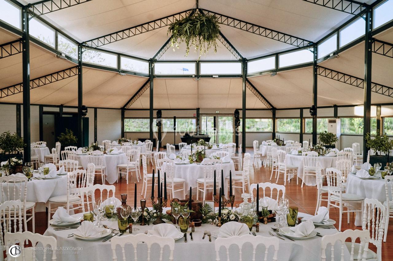 Salle de réception lumineuse avec tables rondes dressées, chaises blanches et décoration végétale suspendue.