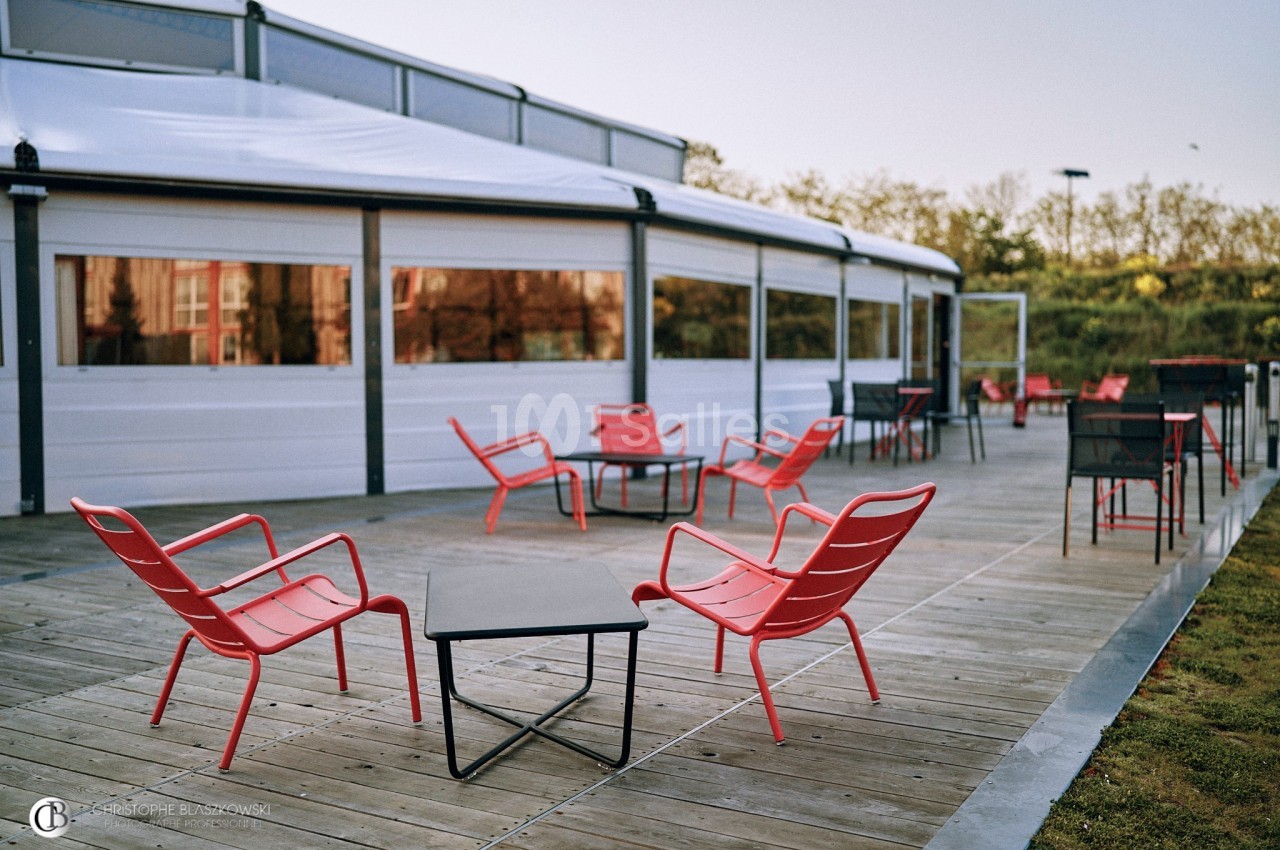 Terrasse en bois avec des tables et chaises rouges disposées autour, proche d'un bâtiment vitré.