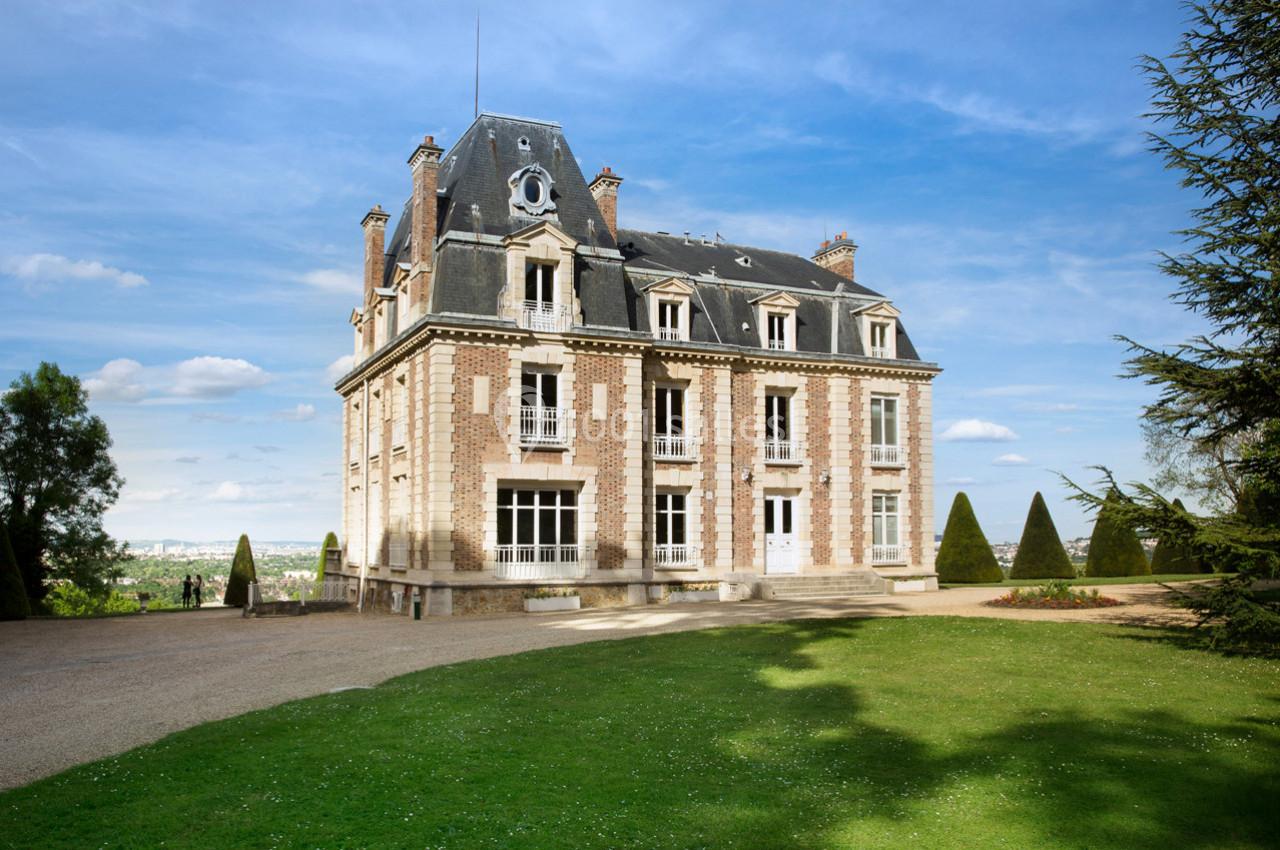 Façade d'un manoir en briques et pierre entouré d'un jardin avec pelouse et arbres, sous un ciel dégagé.