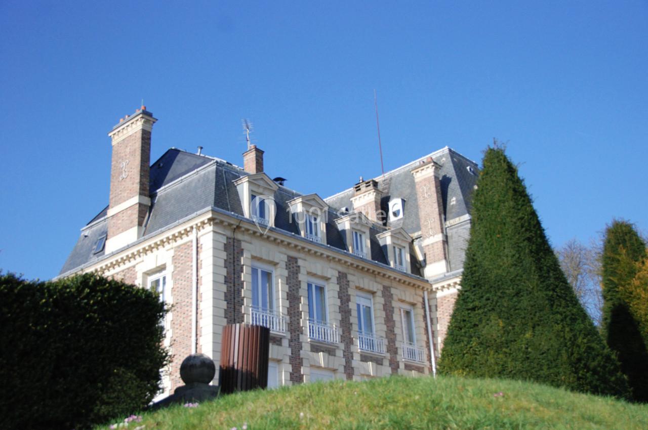 Façade d'un bâtiment en briques et pierres avec toit mansardé, entouré de haies et d'arbres sous un ciel bleu.