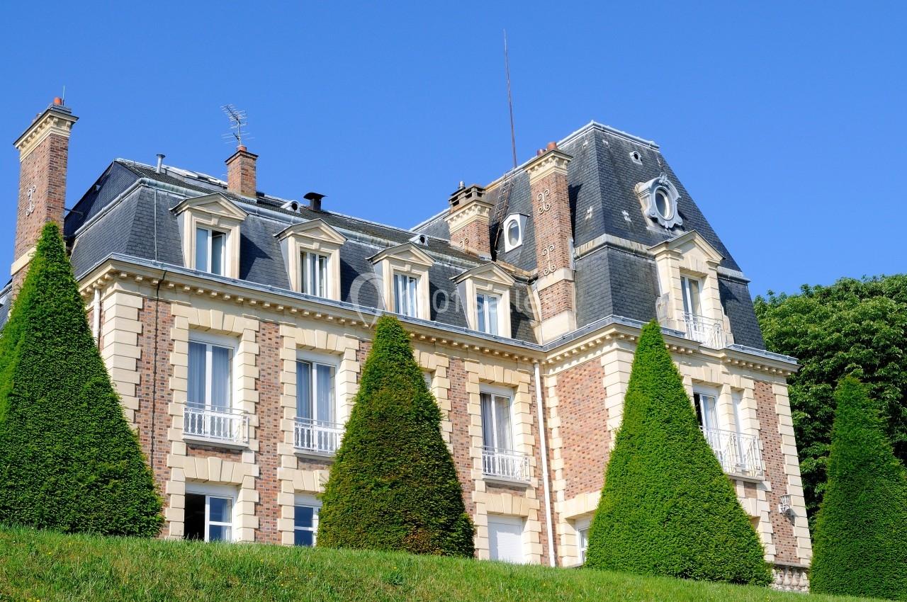 Façade d'un grand bâtiment en briques avec toit mansardé, entouré de conifères taillés et d'une pelouse verte.