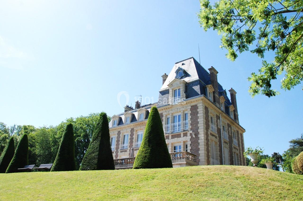 Manoir en pierre entouré d'arbres et de pelouses bien entretenues sous un ciel bleu clair.