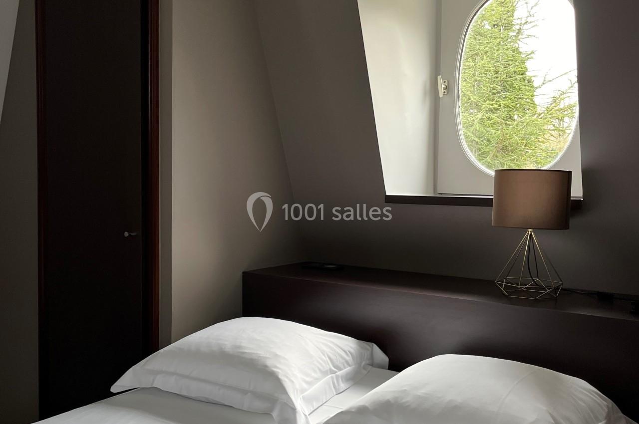 Lit double avec draps blancs dans une chambre mansardée, éclairée par une fenêtre ovale avec vue sur des arbres.