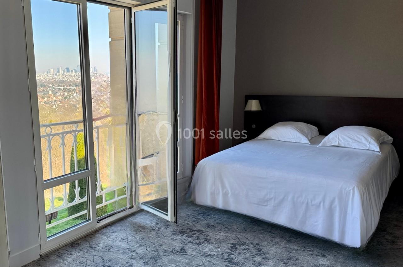 Chambre lumineuse avec lit double, grande fenêtre ouverte sur un balcon offrant une vue sur une ville au loin.