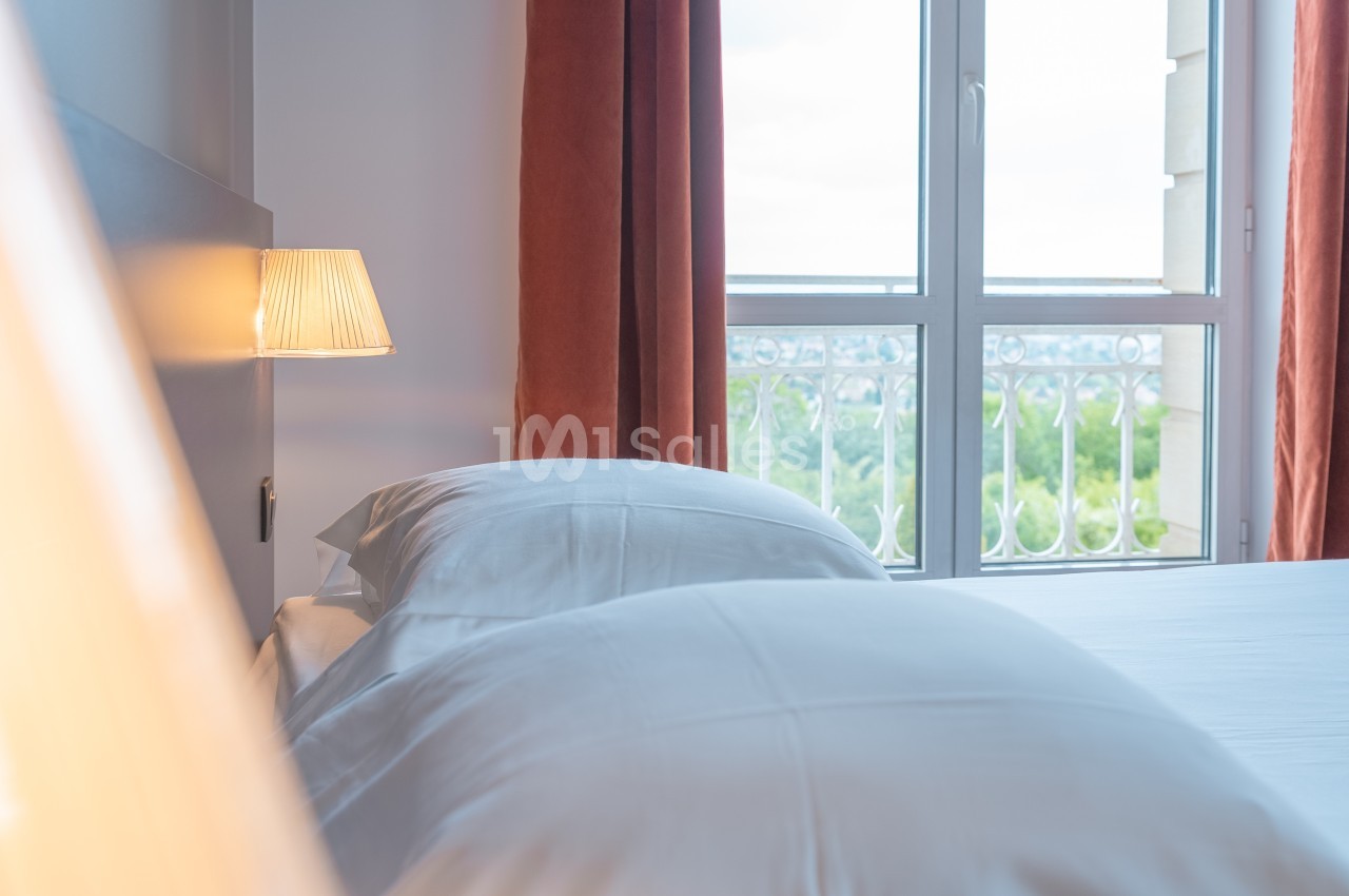 Lit avec draps blancs près d'une lampe allumée, devant une fenêtre donnant sur un paysage verdoyant.