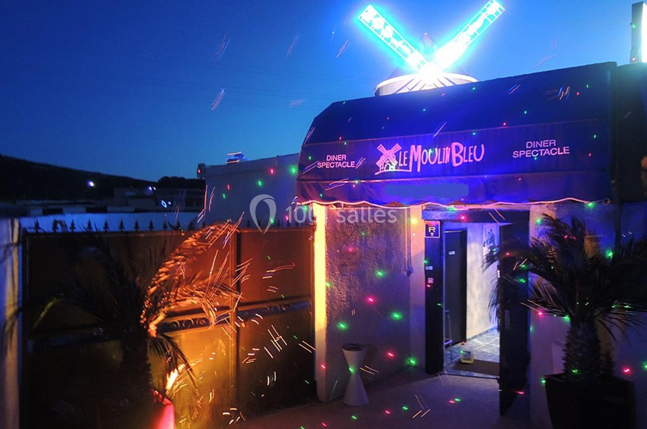 Entrée illuminée d'un cabaret avec enseigne ’Le Moulin Bleu’, décorée de palmiers et lumières colorées.