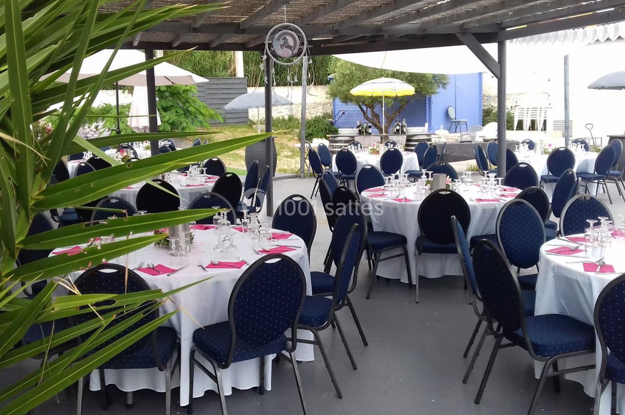 Tables rondes dressées avec nappes blanches et chaises bleues sous une pergola, dans un espace extérieur aménagé.