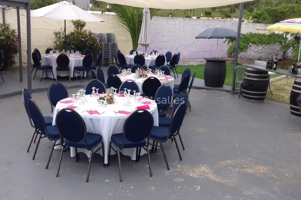 Tables rondes dressées avec nappes blanches, chaises bleues et décorations, installées en extérieur sous des parasols.
