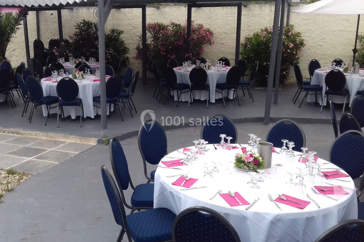 Tables rondes dressées avec nappes blanches, serviettes roses et verres, disposées dans un espace extérieur couvert.