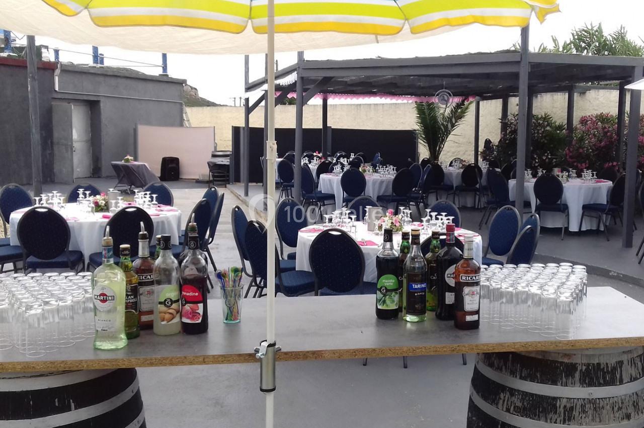 Tables rondes dressées pour un événement en extérieur, avec un bar en premier plan proposant des bouteilles et des verres.