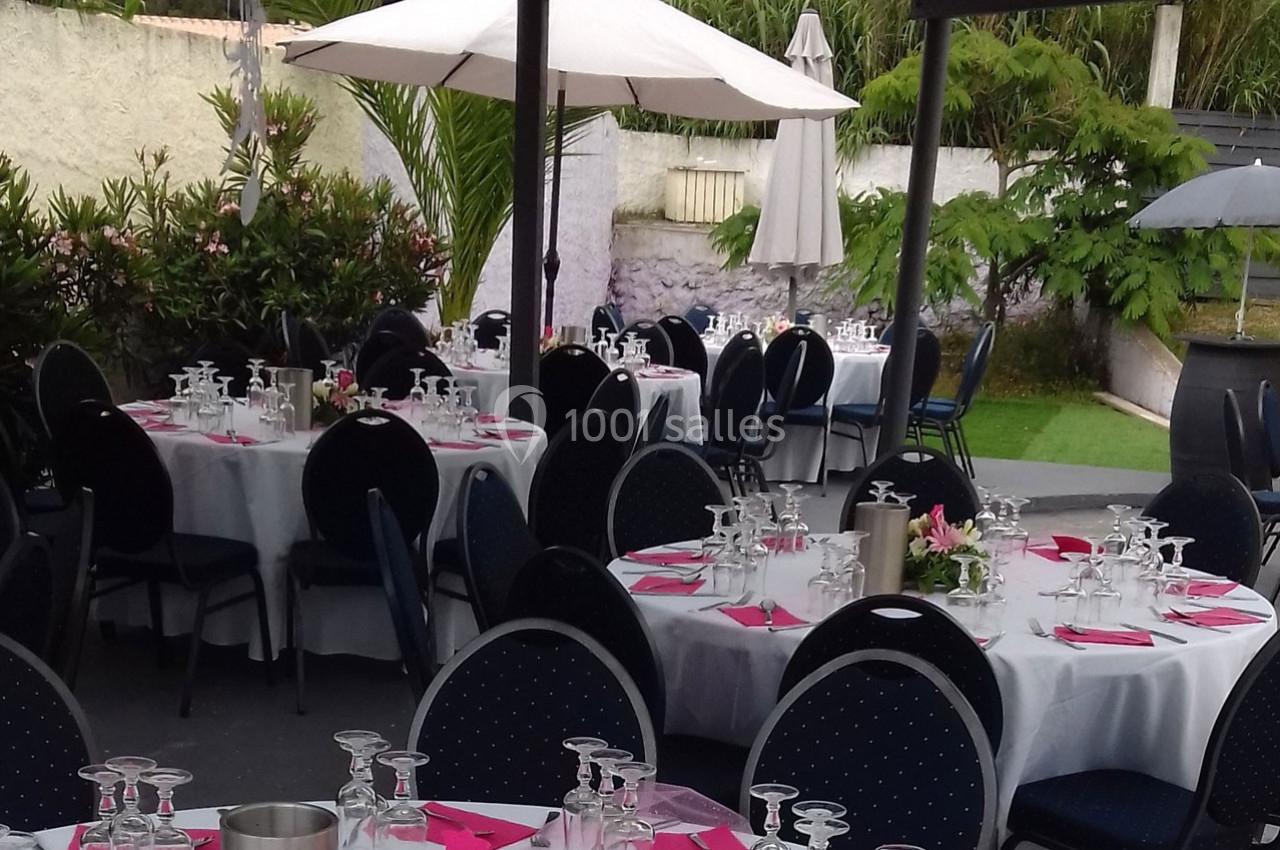 Tables rondes dressées avec nappes blanches, serviettes roses et verres, disposées en extérieur sous des parasols.