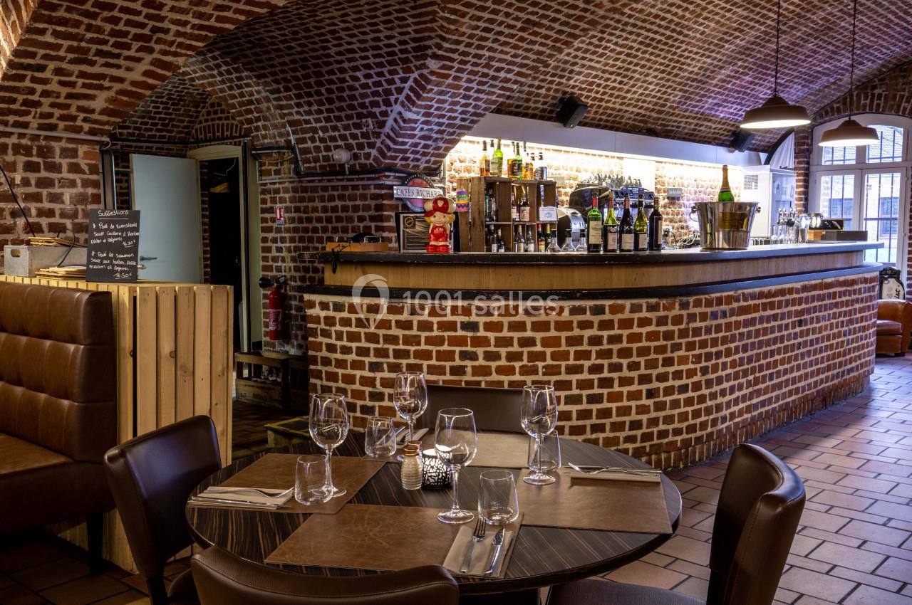 Salle de restaurant avec murs en briques, bar éclairé en arrière-plan et table dressée au premier plan.