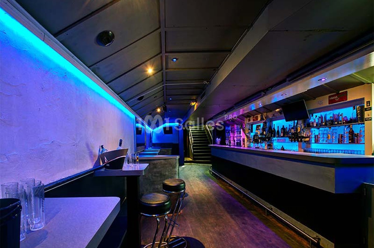 Intérieur d'un bar avec éclairage bleu, comptoir à droite, tables et tabourets à gauche, escalier au fond.