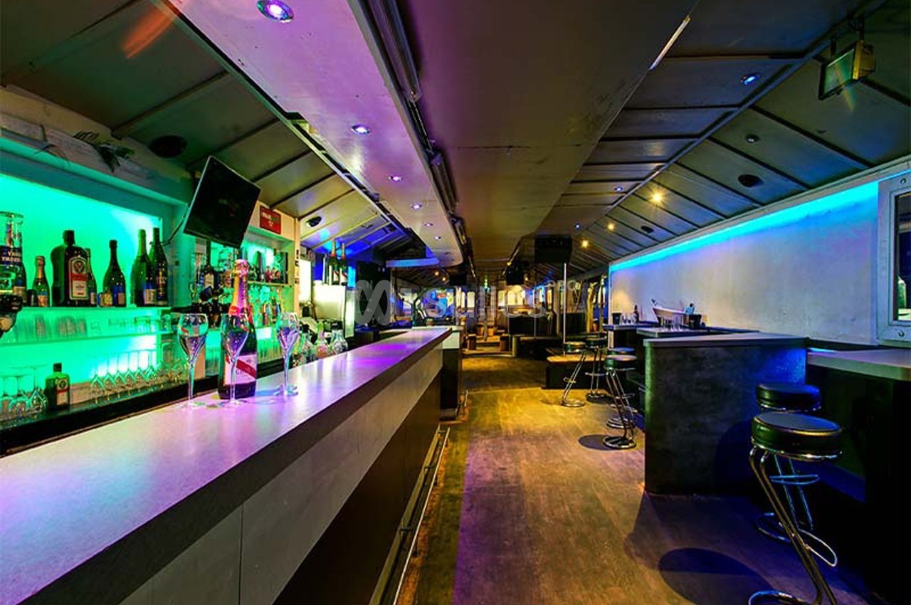 Intérieur d'un bar moderne avec éclairage coloré, comptoirs alignés et tabourets hauts dans une ambiance nocturne.