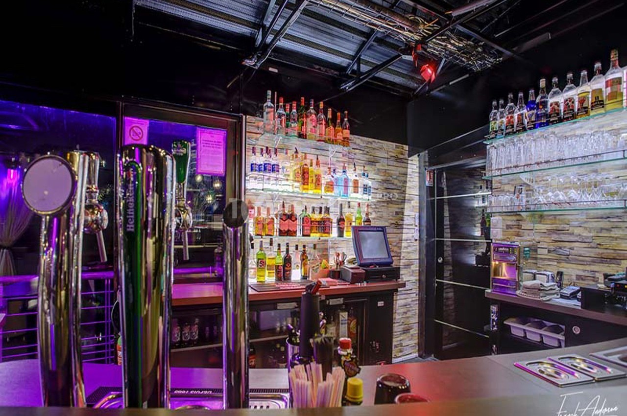 Comptoir de bar moderne avec tireuses à bière, étagères de bouteilles illuminées et caisse enregistreuse.