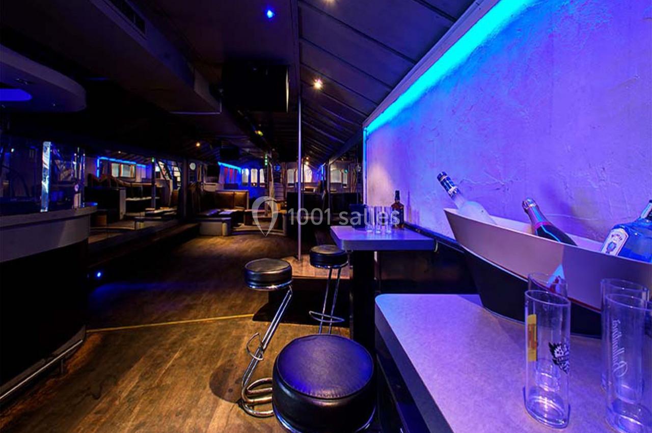 Intérieur d'un bar ou club avec comptoir, tabourets, bouteilles et éclairage bleu tamisé.