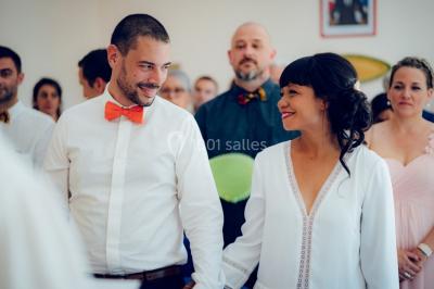 Un couple souriant se tient la main lors d'une cérémonie, entouré d'invités attentifs en arrière-plan.