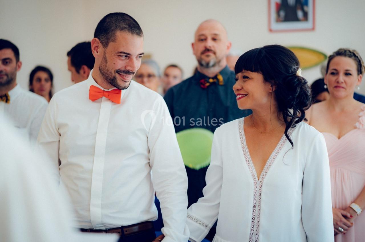 Un couple souriant se tient la main lors d'une cérémonie, entouré d'invités attentifs en arrière-plan.