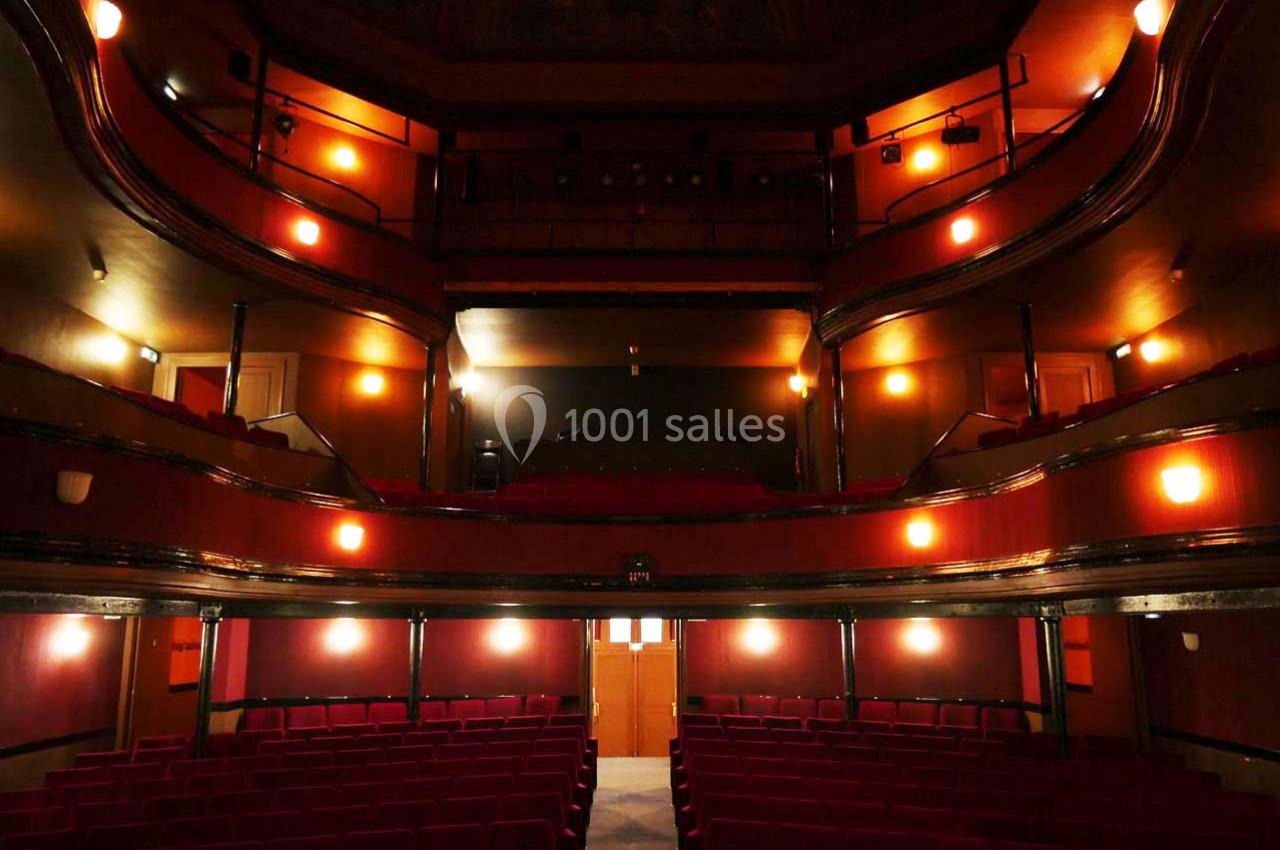 Salle de théâtre vide avec sièges rouges, balcons en bois et éclairage tamisé.
