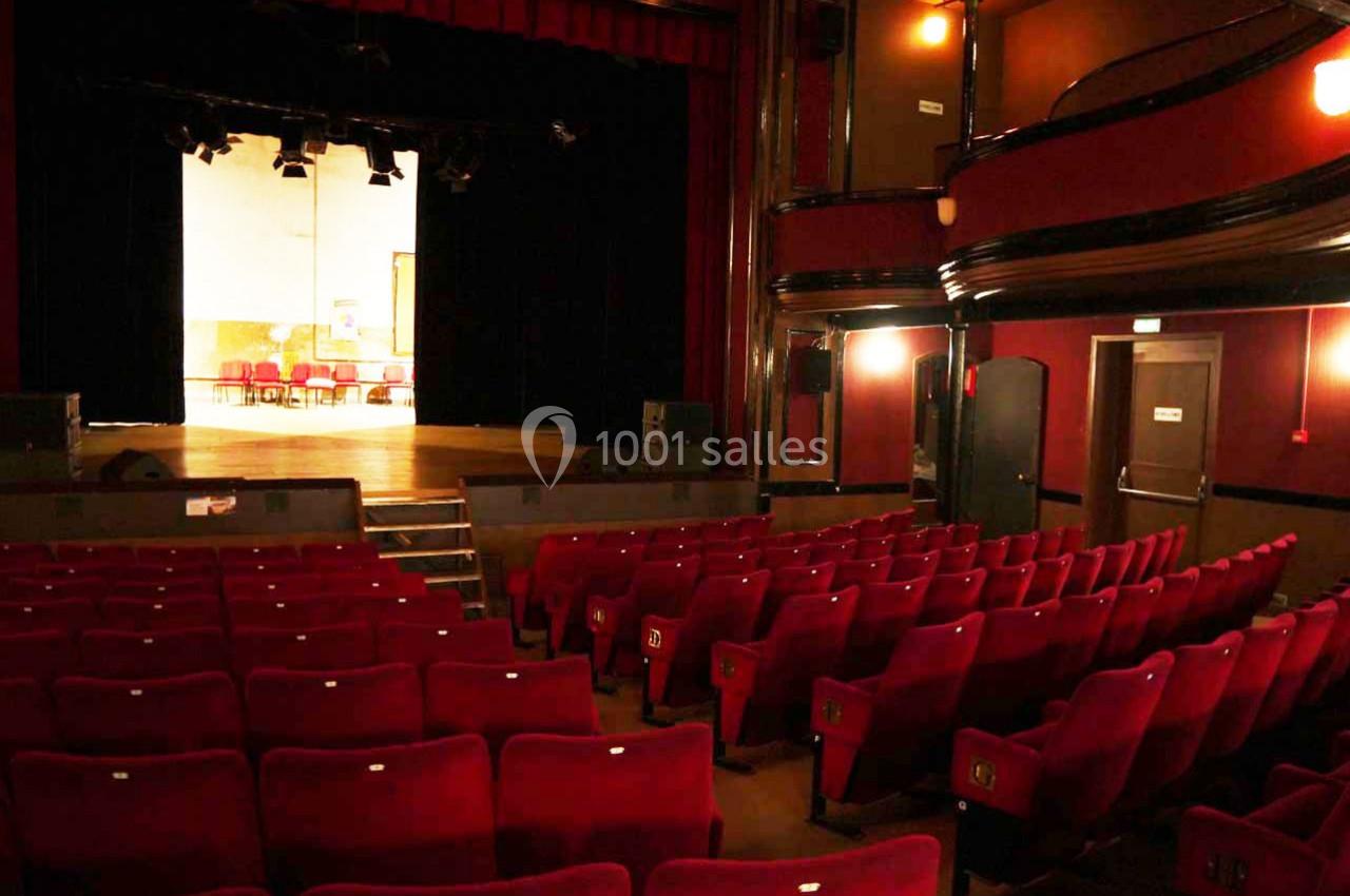 Salle de théâtre vide avec des rangées de fauteuils rouges, une scène éclairée et des murs sombres.