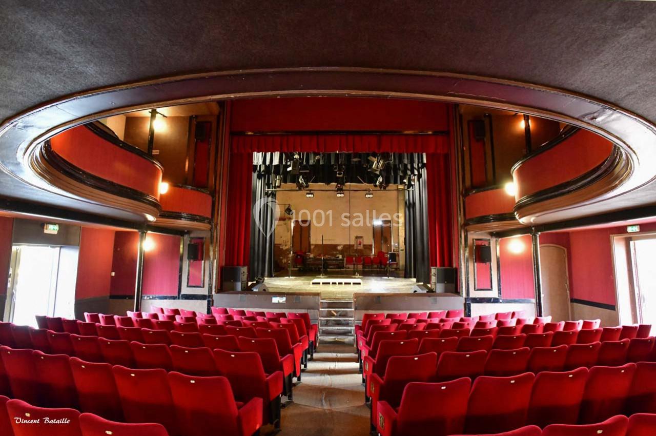 Salle de théâtre vide avec sièges rouges, scène éclairée et rideaux rouges dans un décor classique.