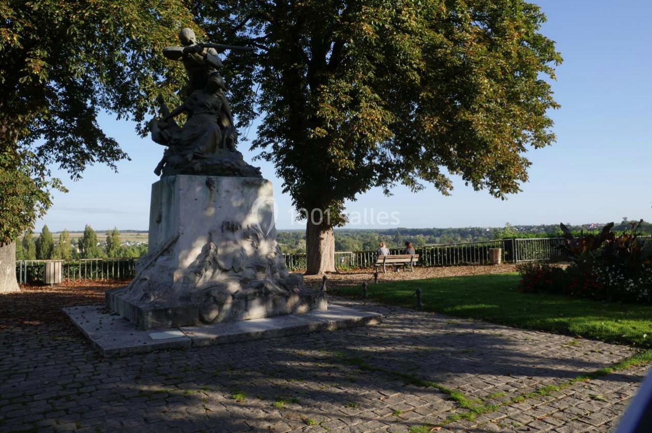 Statue en pierre sur un socle, entourée d'arbres et de verdure, avec une vue dégagée sur un paysage vallonné.