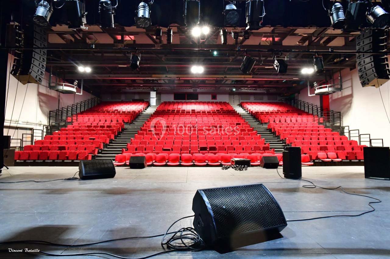 Salle de spectacle vide avec des rangées de sièges rouges, vue depuis la scène équipée de micros et de haut-parleurs.