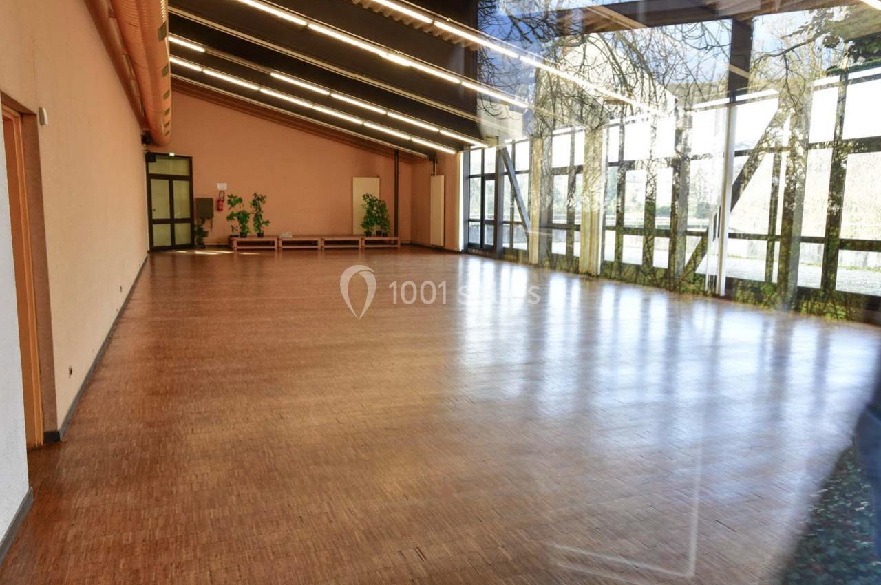 Salle vide avec parquet en bois, grandes baies vitrées laissant entrer la lumière et quelques plantes en décoration.
