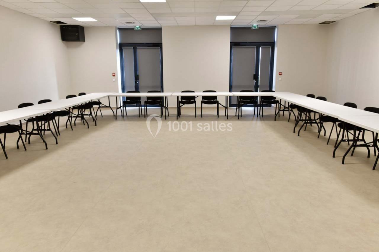 Salle de réunion vide avec des tables blanches disposées en U et des chaises noires, éclairée par des lumières au plafond.