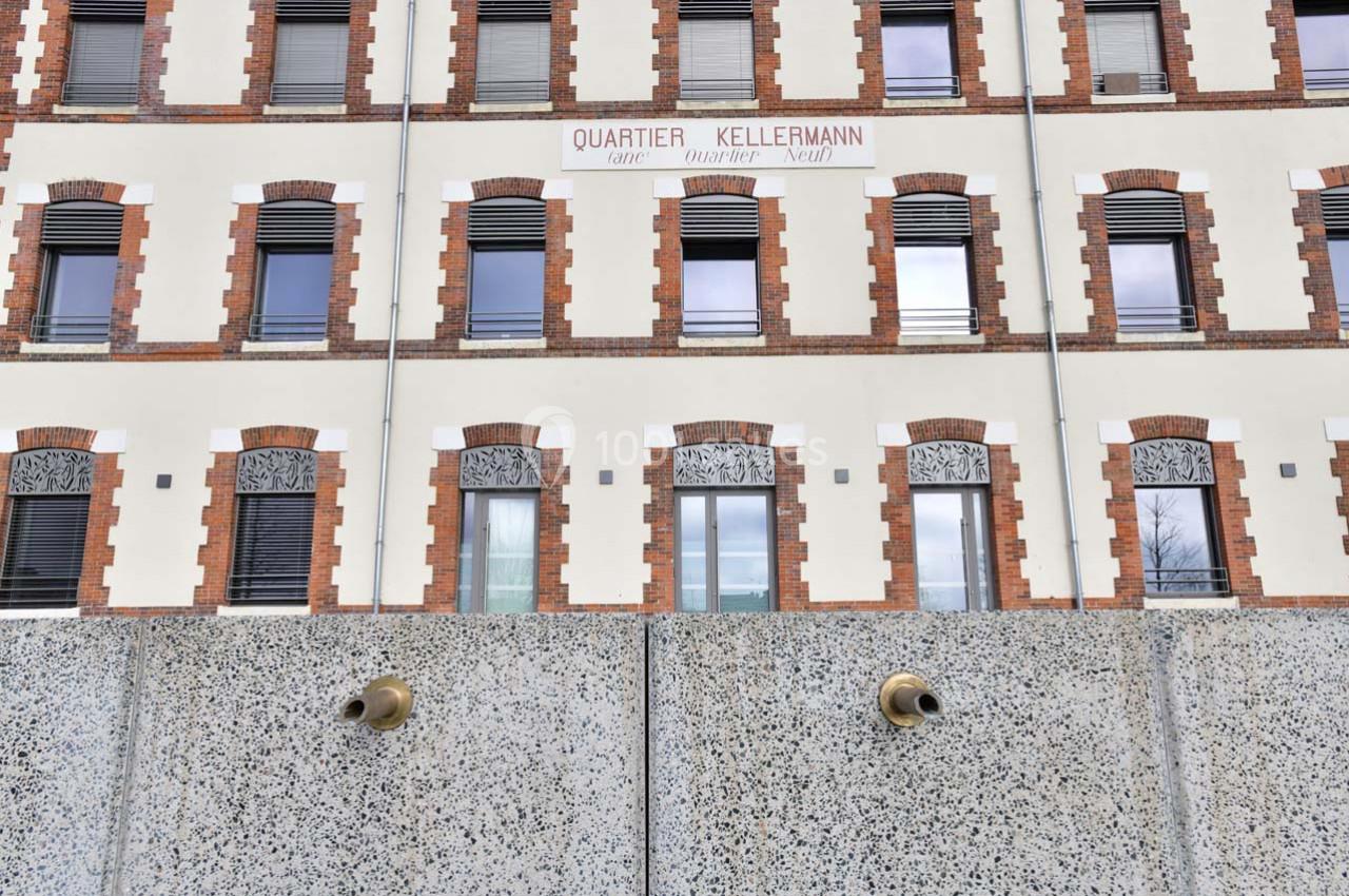 Façade d'un bâtiment en briques et crépi avec des fenêtres alignées, vue derrière un mur en béton percé de tuyaux.