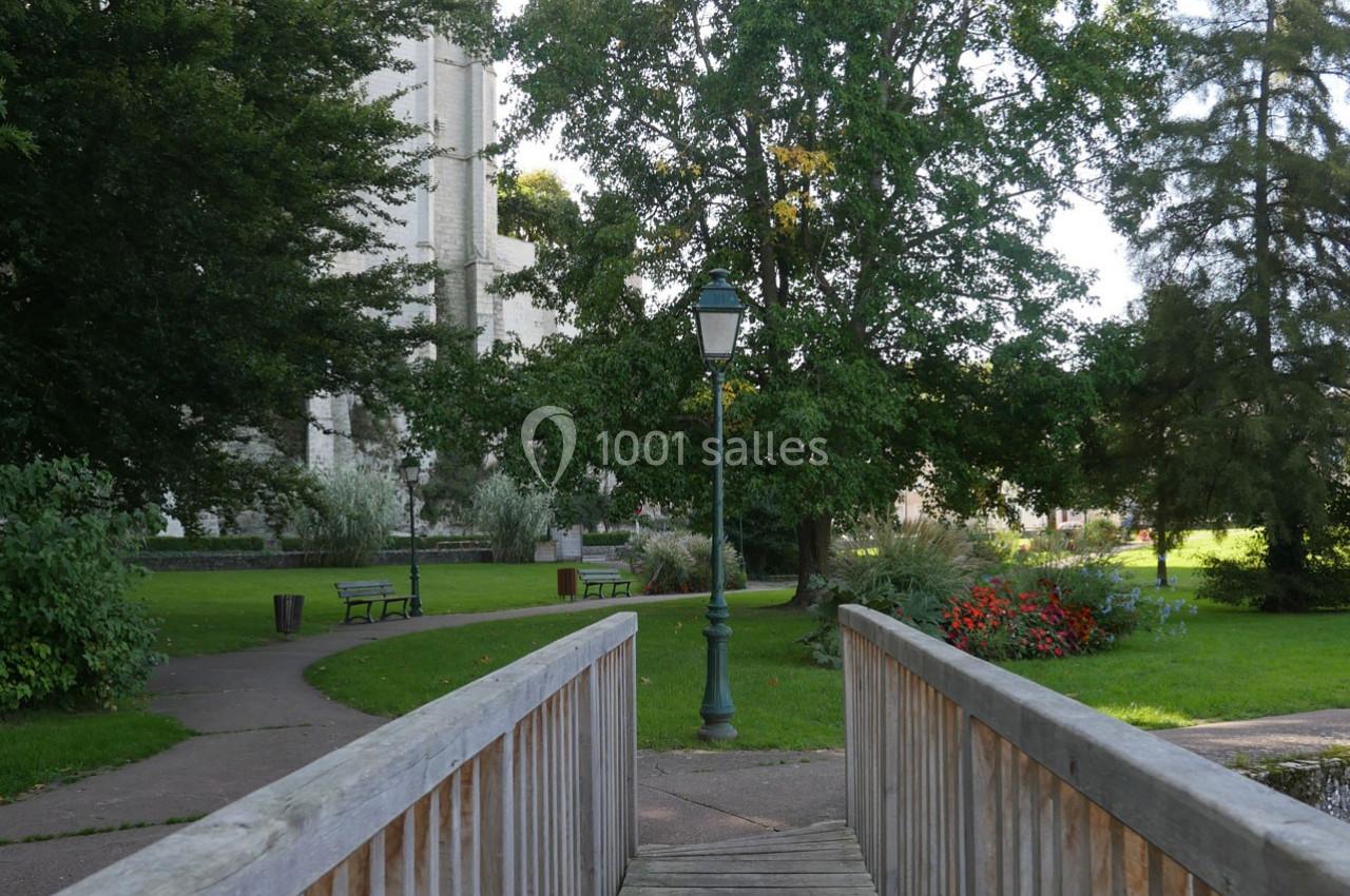 Passerelle en bois menant à un parc verdoyant avec lampadaire, bancs et arbres en arrière-plan.