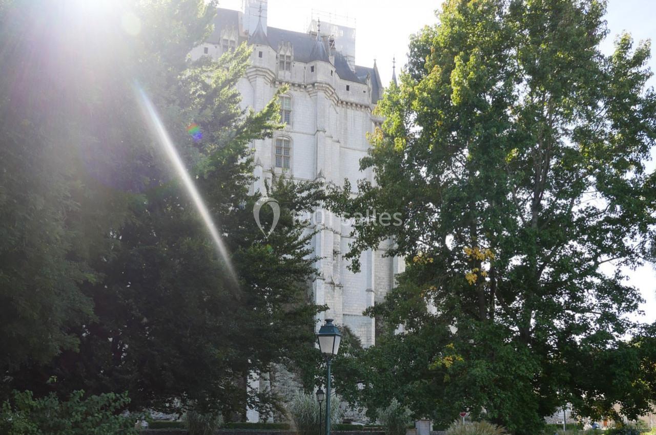 Façade d'un bâtiment historique en pierre blanche partiellement cachée par des arbres, sous une lumière ensoleillée.