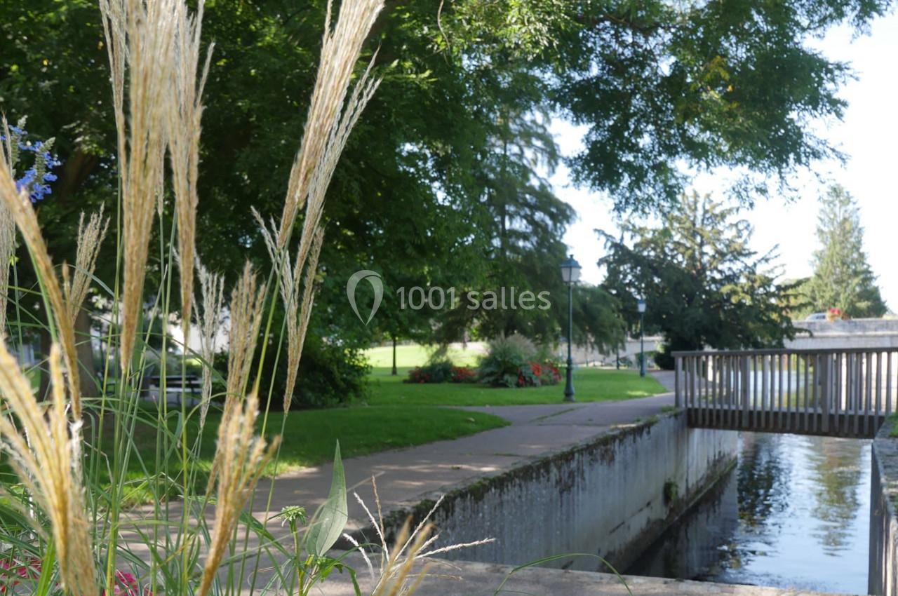 Herbes hautes au premier plan près d'un canal bordé d'arbres et d'un petit pont en bois dans un parc.