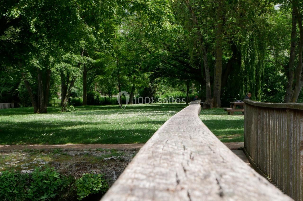 Vue d'un parc verdoyant avec un pont en bois au premier plan et des arbres ombrageant une pelouse.