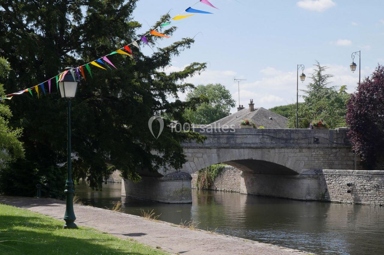 Pont en pierre traversant une rivière bordée d'arbres, avec des fanions colorés suspendus et un lampadaire au premier plan.