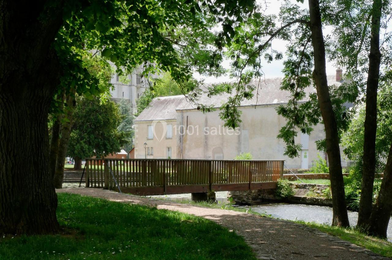 Petit pont en bois traversant un cours d'eau, entouré d'arbres et d'herbe, avec des bâtiments en arrière-plan.