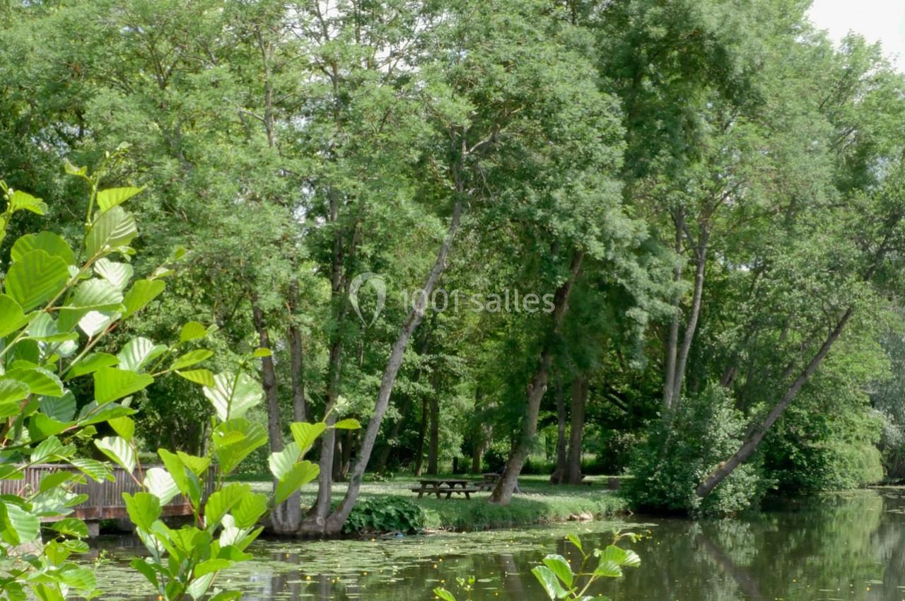 Bord d’un étang entouré d’arbres avec une table de pique-nique visible sur la rive herbeuse.
