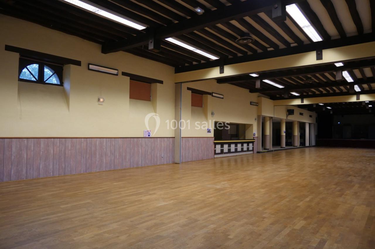 Salle vide avec parquet en bois, murs jaunes, poutres apparentes au plafond et fenêtres en hauteur.
