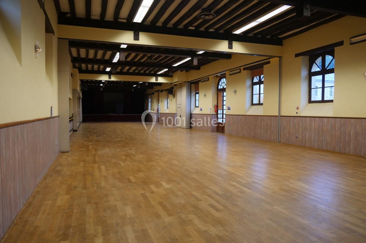 Salle vide avec parquet en bois, murs jaunes, poutres apparentes au plafond et grandes fenêtres en arc.