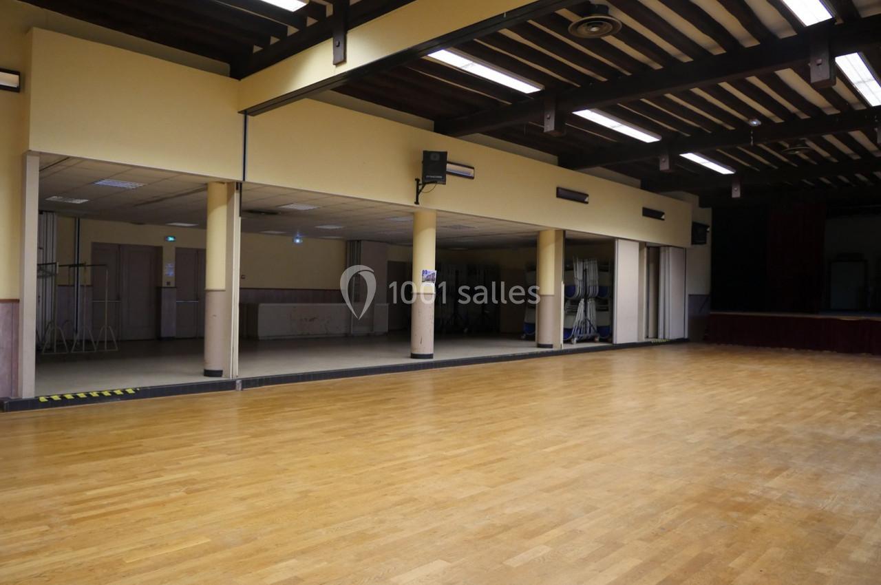 Salle spacieuse avec parquet en bois, poutres apparentes au plafond et scène en arrière-plan.