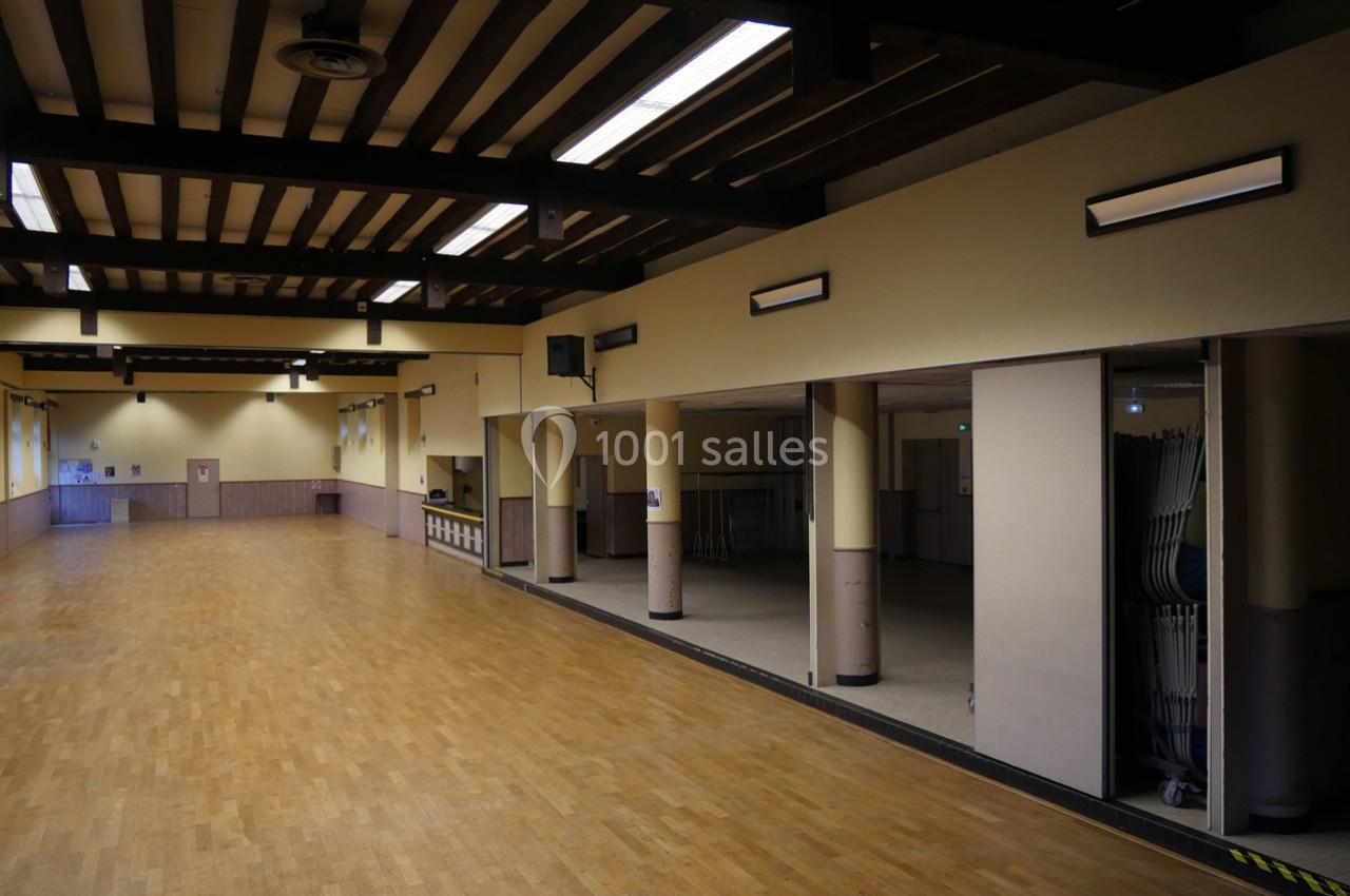 Grande salle vide avec parquet au sol, murs clairs, poutres apparentes au plafond et éclairage fluorescent.