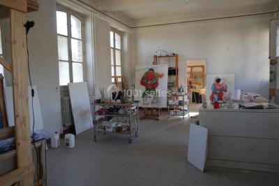 Atelier d'artiste lumineux avec tables de travail, matériel de peinture, étagères et une toile en cours.