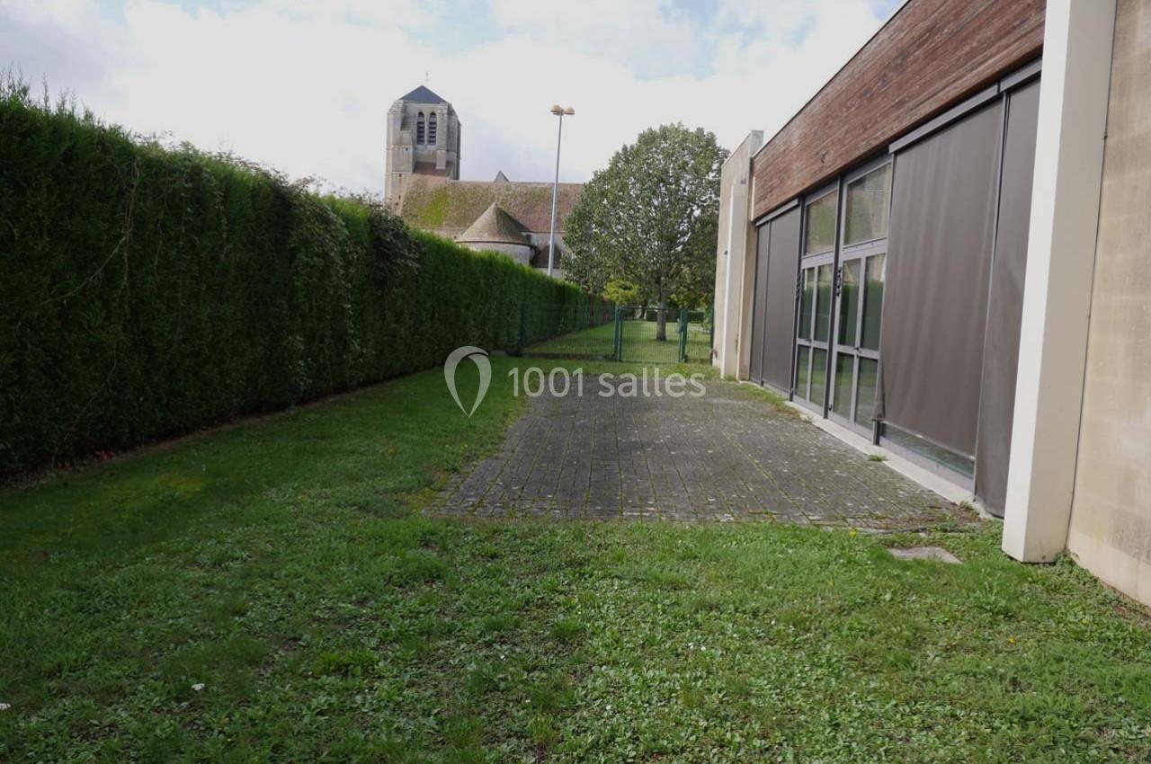 Allée enherbée bordée de haies et d'un bâtiment moderne, avec une église en arrière-plan sous un ciel dégagé.