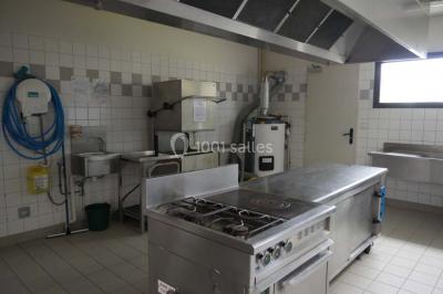 Cuisine professionnelle avec plan de travail en inox, cuisinière, évier, hotte et équipements muraux dans un espace carrelé.